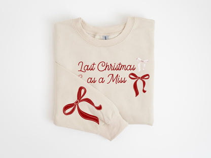 May include: Cream-colored sweatshirt with the text "Last Christmas as a Miss" in red script. The design includes red and white bow illustrations. A festive holiday-themed garment.