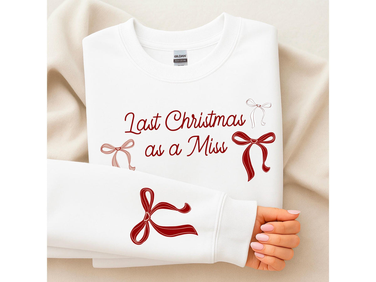 May include: White sweatshirt with the text "Last Christmas as a Miss" in burgundy script. The design includes several bow illustrations in burgundy and blush pink. The sleeve of the sweatshirt is visible, with a large bow graphic.