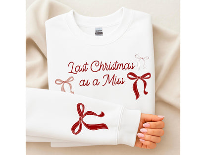 May include: White sweatshirt with the text "Last Christmas as a Miss" in burgundy script. The design includes several bow illustrations in burgundy and blush pink. The sleeve of the sweatshirt is visible, with a large bow graphic.