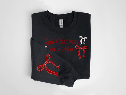 May include: Black sweatshirt with the text "Last Christmas as a Miss" in red script. The sweatshirt features red and white bow designs. The folded garment is displayed on a white background.