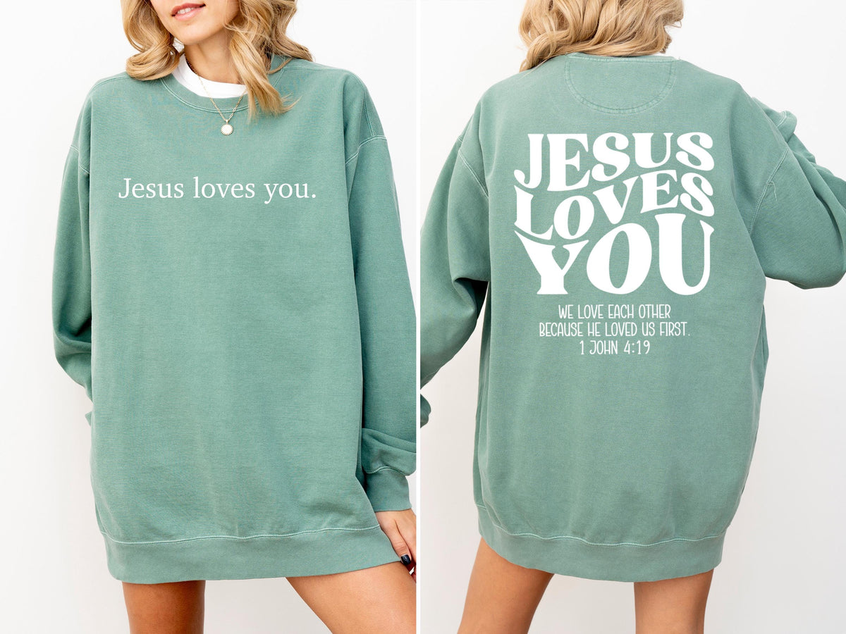 May include: A sage green sweatshirt with the text "Jesus loves you" on the front. The back of the sweatshirt features the words "Jesus Loves You" in a stylized font, along with a Bible verse.