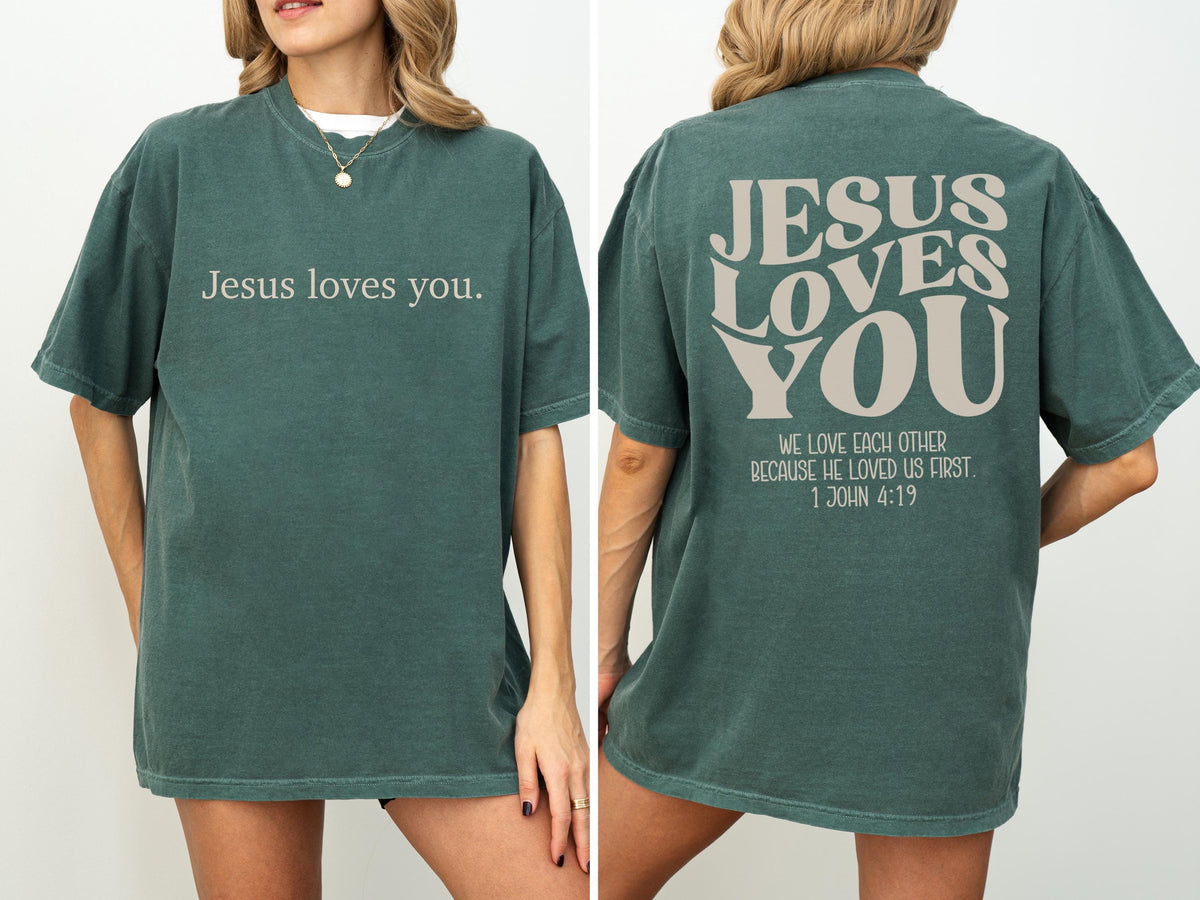 May include: Dark teal t-shirt with the text "Jesus loves you." on the front. The back of the shirt features the words "JESUS LOVES YOU" and the quote "WE LOVE EACH OTHER BECAUSE HE LOVED US FIRST. 1 JOHN 4:19."