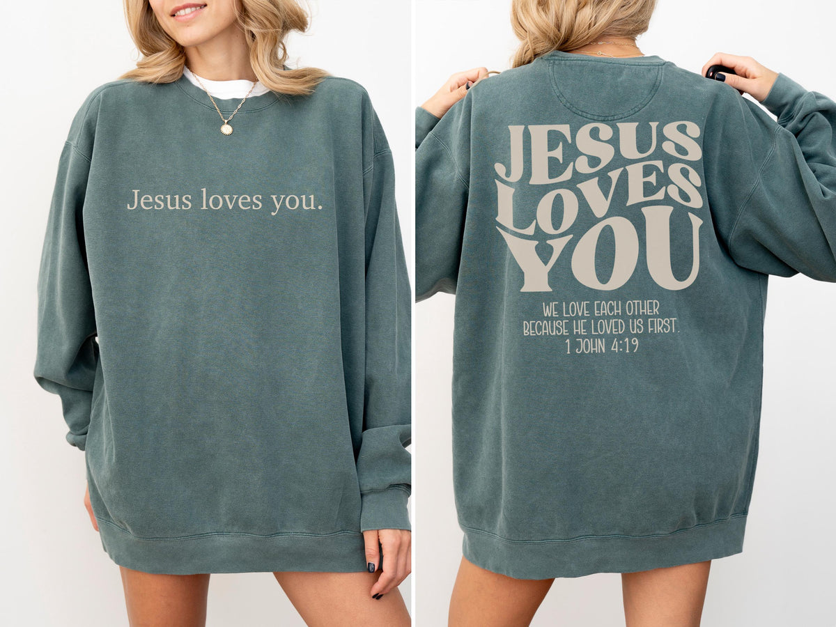 May include: A teal-colored sweatshirt with the text "Jesus loves you" on the front. The back of the sweatshirt features the words "JESUS LOVES YOU" and the quote "WE LOVE EACH OTHER BECAUSE HE LOVED US FIRST 1 JOHN 4:19".