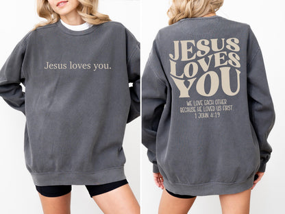 May include: A charcoal gray sweatshirt with the text "Jesus loves you" on the front. The back of the sweatshirt features the words "JESUS LOVES YOU" in a stylized font, along with the quote "WE LOVE EACH OTHER BECAUSE HE LOVED US FIRST 1 JOHN 4:19."