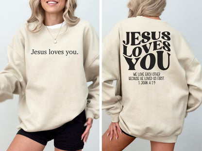 May include: Beige sweatshirt with the text "Jesus loves you" on the front. The back of the sweatshirt features the words "Jesus Loves You" in a bold, retro font, along with the Bible verse 1 John 4:19.
