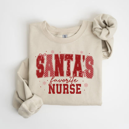 May include: A beige sweatshirt with the text "SANTA'S favorite NURSE" in red, checkered letters. The design includes red bows and snowflakes. The sleeves are folded, and a matching scrunchie is visible.