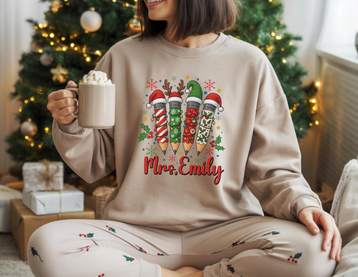 May include: Beige sweatshirt featuring a Christmas-themed pencil design with Santa hats and festive patterns. The text "Mrs. Emily" is displayed in red script. The person is holding a beige mug with whipped cream.