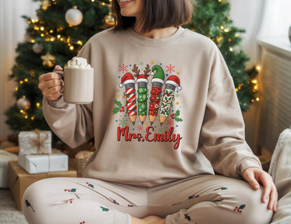 May include: Beige sweatshirt featuring a Christmas-themed pencil design with Santa hats and festive patterns. The text "Mrs. Emily" is displayed in red script. The person is holding a beige mug with whipped cream.
