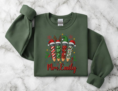 May include: A dark green sweatshirt featuring a festive design with four pencils wearing Christmas hats. The pencils are decorated with holiday patterns and the name "Mrs. Emily" in red script.