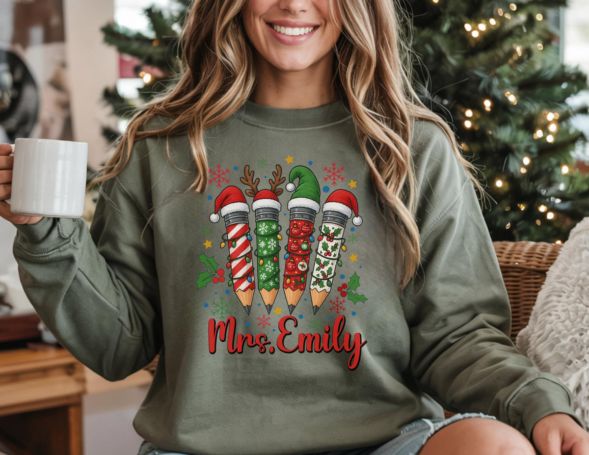 May include: Olive green sweatshirt featuring a festive pencil design with Santa hats and holiday decorations. The pencils are decorated with red and white stripes, green holly, and snowflakes. The name "Mrs. Emily" is written in red script below the pencils.