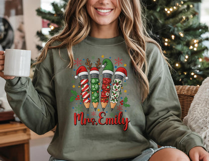 May include: Olive green sweatshirt featuring a festive pencil design with Santa hats and holiday decorations. The pencils are decorated with red and white stripes, green holly, and snowflakes. The name "Mrs. Emily" is written in red script below the pencils.