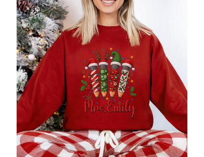May include: Red sweatshirt featuring a Christmas pencil design with festive hats and holly. The pencils are decorated with holiday patterns. The name "Mrs. Emily" is written in cursive below the pencils. The model is wearing red and white plaid pajama pants.