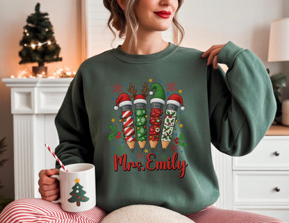 May include: A teal green sweatshirt featuring a festive pencil design with Santa hats and holiday decorations. The pencils are decorated with stripes, lights, and holly. The name "Mrs. Emily" is written in red script below the pencils. A white mug with a Christmas tree design is held in the hand.