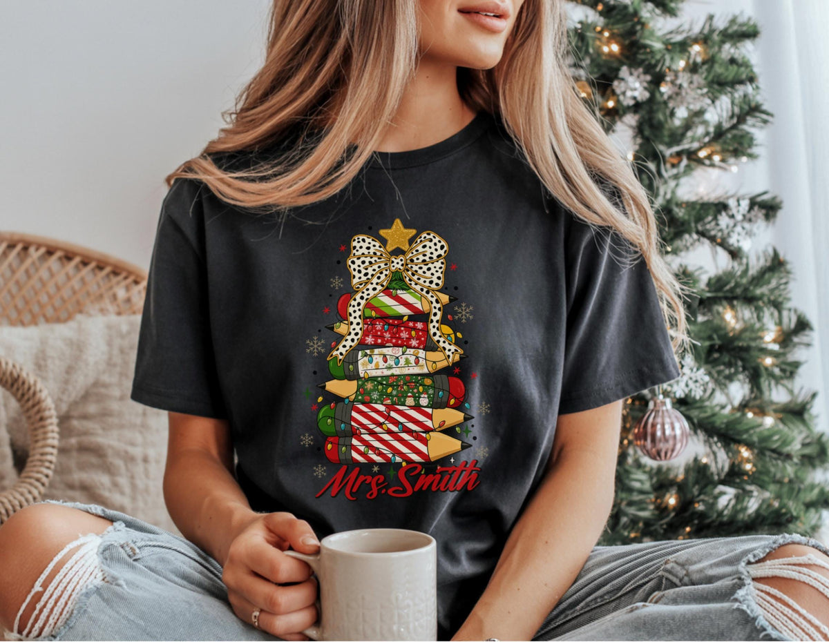 May include: Dark gray t-shirt featuring a Christmas tree design made of wrapped gifts. The tree is topped with a gold star and a leopard print bow. The name "Mrs. Smith" is written in red script below the tree.