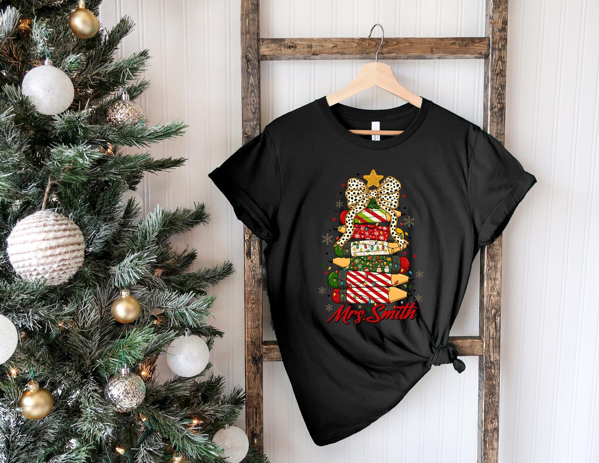 May include: Black t-shirt featuring a Christmas tree design made of stacked gift boxes. The tree has a leopard print bow, a gold star, and the text "Mrs. Smith" in red. The shirt is hanging on a wooden ladder.