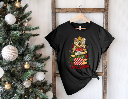 May include: Black t-shirt featuring a Christmas tree design made of stacked gift boxes. The tree has a leopard print bow, a gold star, and the text "Mrs. Smith" in red. The shirt is hanging on a wooden ladder.