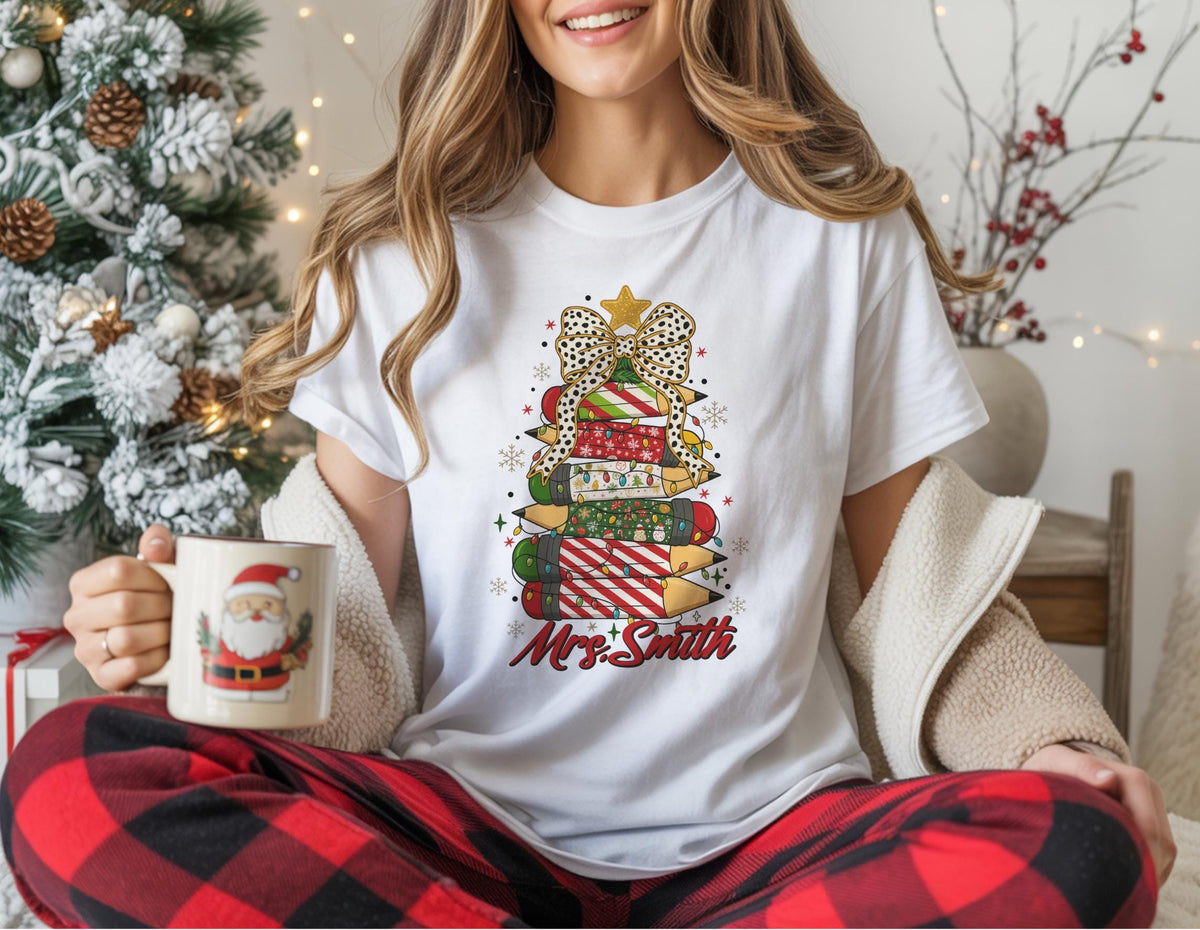 May include: White t-shirt featuring a Christmas tree design made of pencils, bows, and presents. The design includes a gold star, snowflakes, and the name "Mrs. Smith" in red script. The person is holding a Santa mug.
