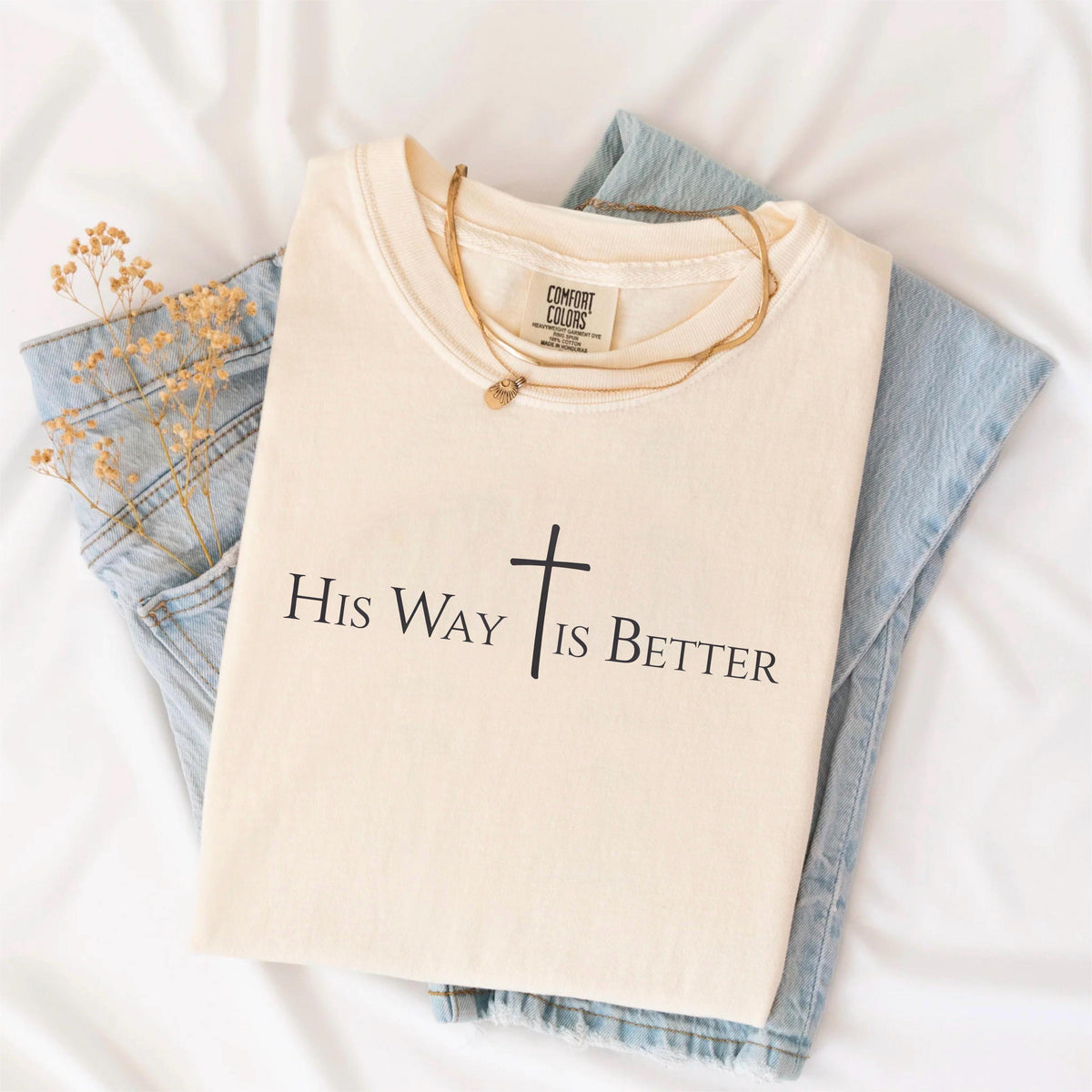 May include: Cream-colored t-shirt with the text "His Way Is Better" and a cross graphic in black. The shirt is folded and placed on top of a pair of light blue jeans. A gold necklace is draped across the shirt.