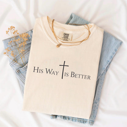 May include: Cream-colored t-shirt with the text "His Way Is Better" and a cross graphic in black. The shirt is folded and placed on top of a pair of light blue jeans. A gold necklace is draped across the shirt.