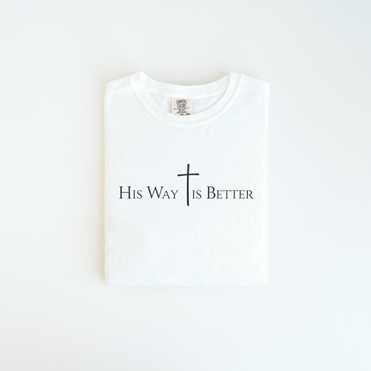 May include: A folded white t-shirt with the text "His Way Is Better" in black, with a cross symbol. The shirt is made of a soft material and is neatly folded, ready to wear.