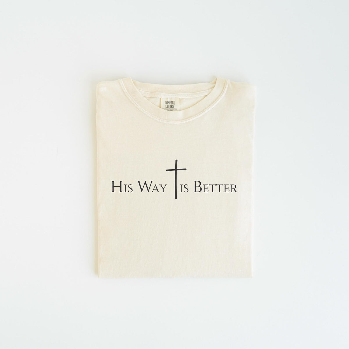 May include: Cream-colored t-shirt with the text "His Way Is Better" and a cross symbol printed in black. The shirt is folded neatly, showcasing the design prominently. The t-shirt is made of a soft material.