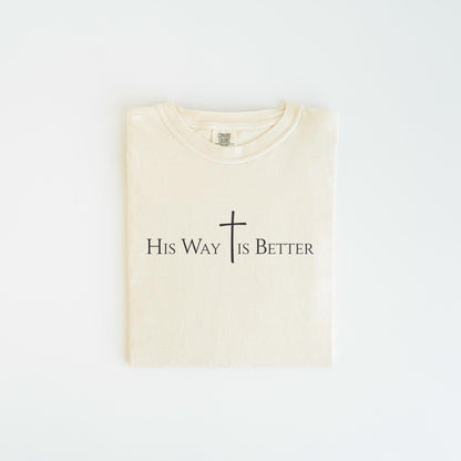 May include: Cream-colored t-shirt with the text "His Way Is Better" and a cross symbol printed in black. The shirt is folded neatly, showcasing the design prominently. The t-shirt is made of a soft material.