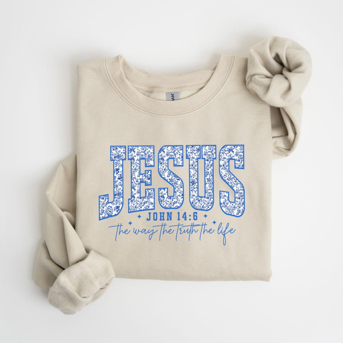 May include: A beige sweatshirt with the word "JESUS" in blue floral lettering. Below the word "JESUS" is the text "JOHN 14:6" and "The way the truth the life" in blue script. The sweatshirt has long sleeves.