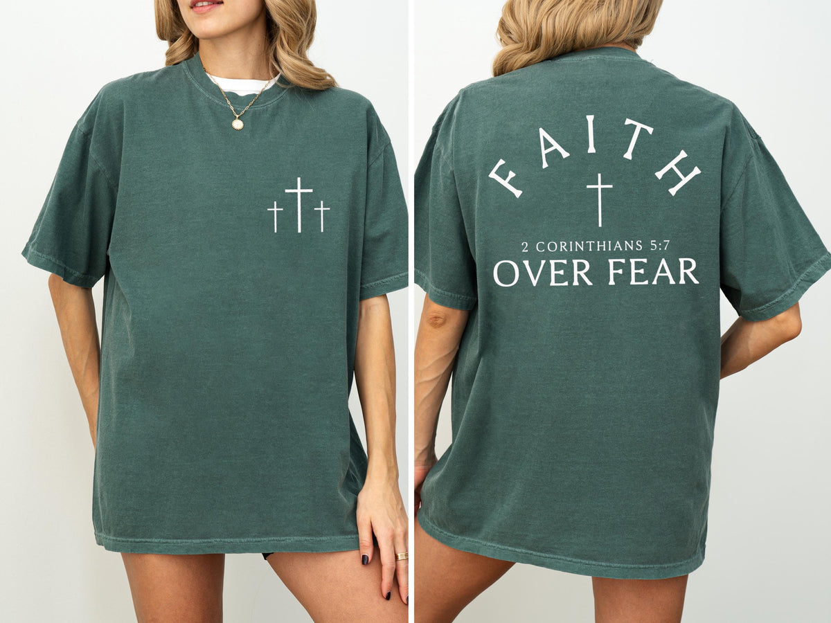 May include: A dark teal t-shirt with white text and graphics. The front features three crosses. The back displays the words "FAITH" above a cross, with "2 CORINTHIANS 5:7 OVER FEAR" below. The shirt has short sleeves and a relaxed fit.