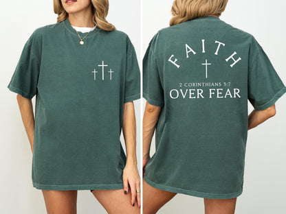 May include: A dark teal t-shirt with white text and graphics. The front features three crosses. The back displays the words "FAITH" above a cross, with "2 CORINTHIANS 5:7 OVER FEAR" below. The shirt has short sleeves and a relaxed fit.