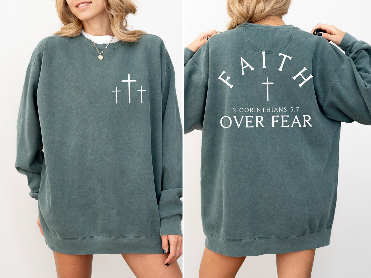 May include: A teal-colored sweatshirt with white text and graphics. The front features three crosses, and the back displays the word "FAITH" above a cross, with "2 CORINTHIANS 5:7 OVER FEAR" below. The sweatshirt has a crew neck.
