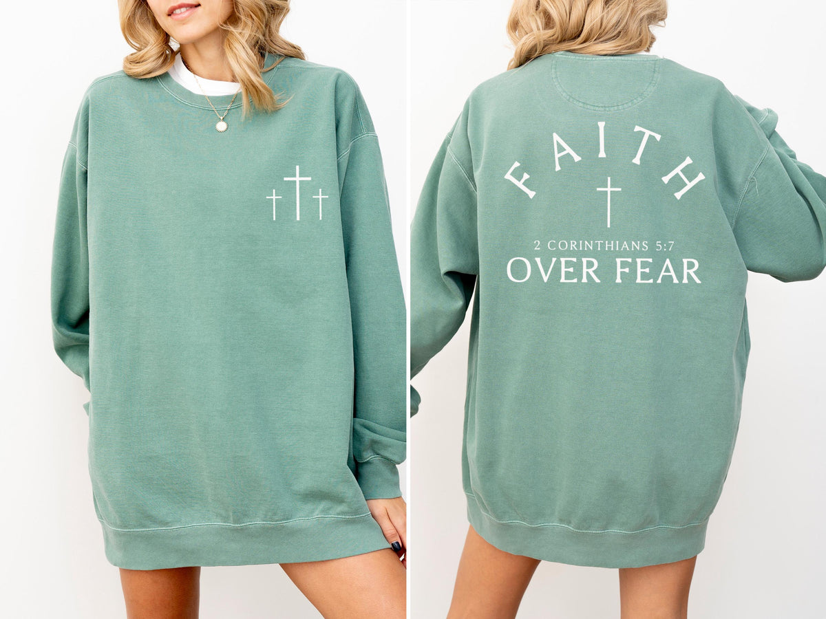 May include: A sage green sweatshirt with white text and cross graphics. The front features three crosses, while the back displays "FAITH" above a cross, with "2 CORINTHIANS 5:7 OVER FEAR" below. The sweatshirt has a crew neck and long sleeves.