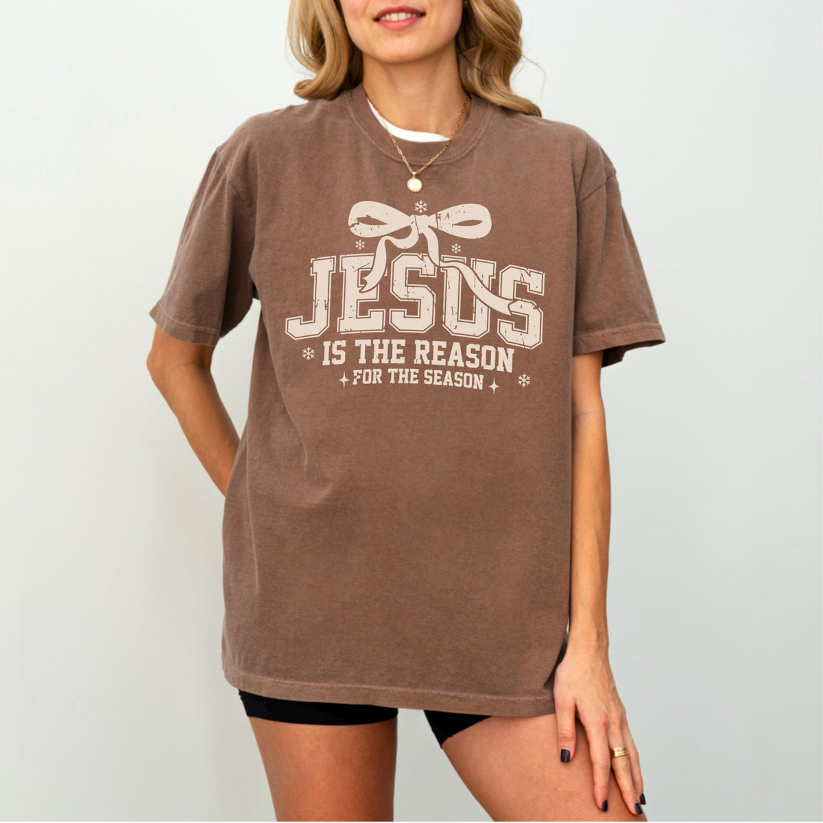 May include: Brown t-shirt with the text "JESUS is the reason for the season" in a cream-colored font. The text is centered on the front of the shirt, with a bow graphic above the word "JESUS". The shirt has short sleeves.