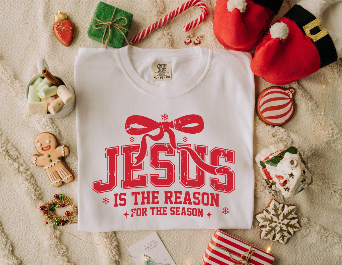 May include: White t-shirt with the red text "JESUS IS THE REASON FOR THE SEASON" and a bow graphic. The shirt is surrounded by Christmas-themed decorations, including cookies, gifts, and Santa hats.