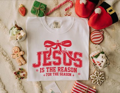 May include: White t-shirt with the red text "JESUS IS THE REASON FOR THE SEASON" and a bow graphic. The shirt is surrounded by Christmas-themed decorations, including cookies, gifts, and Santa hats.