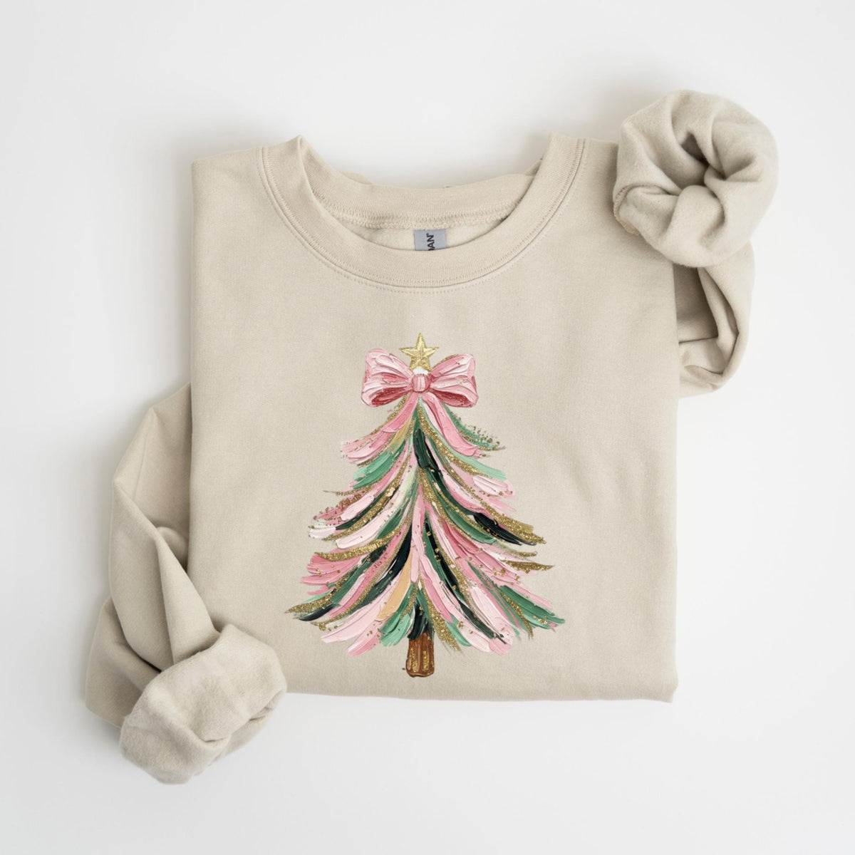 May include: A beige sweatshirt featuring a Christmas tree design. The tree is decorated with pink, green, black, and gold brushstrokes, topped with a pink bow and a gold star. A matching scrunchie rests on the sleeve.