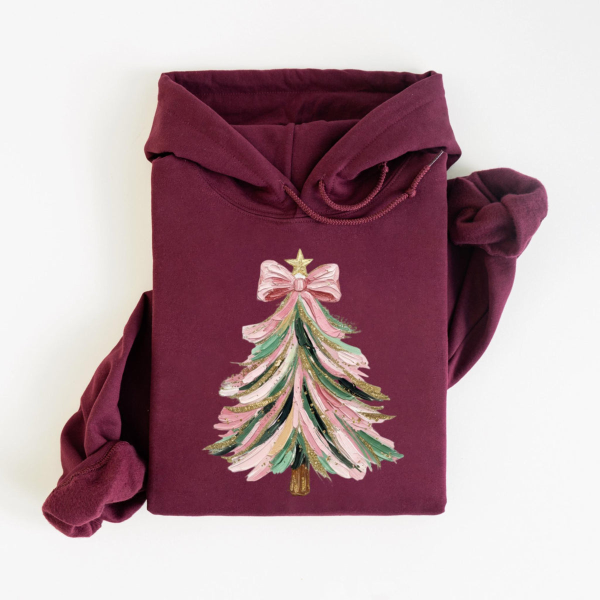 May include: A maroon hoodie featuring a Christmas tree design. The tree is decorated with pink, green, and gold brushstrokes, topped with a pink bow and a gold star. The sleeves are rolled up, and the hood is visible.