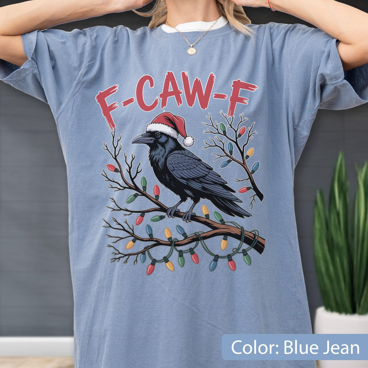 May include: A blue jean colored t-shirt featuring a graphic of a black crow wearing a red and white Santa hat perched on a bare tree branch wrapped in colorful Christmas lights. The text F-CAW-F is printed in red above the crow illustration.