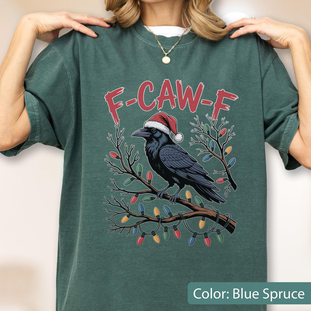 May include: A dark green t-shirt featuring a graphic of a black raven wearing a red and white Santa hat, perched on a brown tree branch wrapped in colorful Christmas lights. The text F-CAW-F is printed in red above the raven illustration.