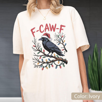 May include: An ivory short-sleeve t-shirt featuring a graphic of a black raven wearing a red and white Santa hat, perched on a bare tree branch wrapped in colorful Christmas lights, with the red text F-CAW-F printed above the bird illustration.