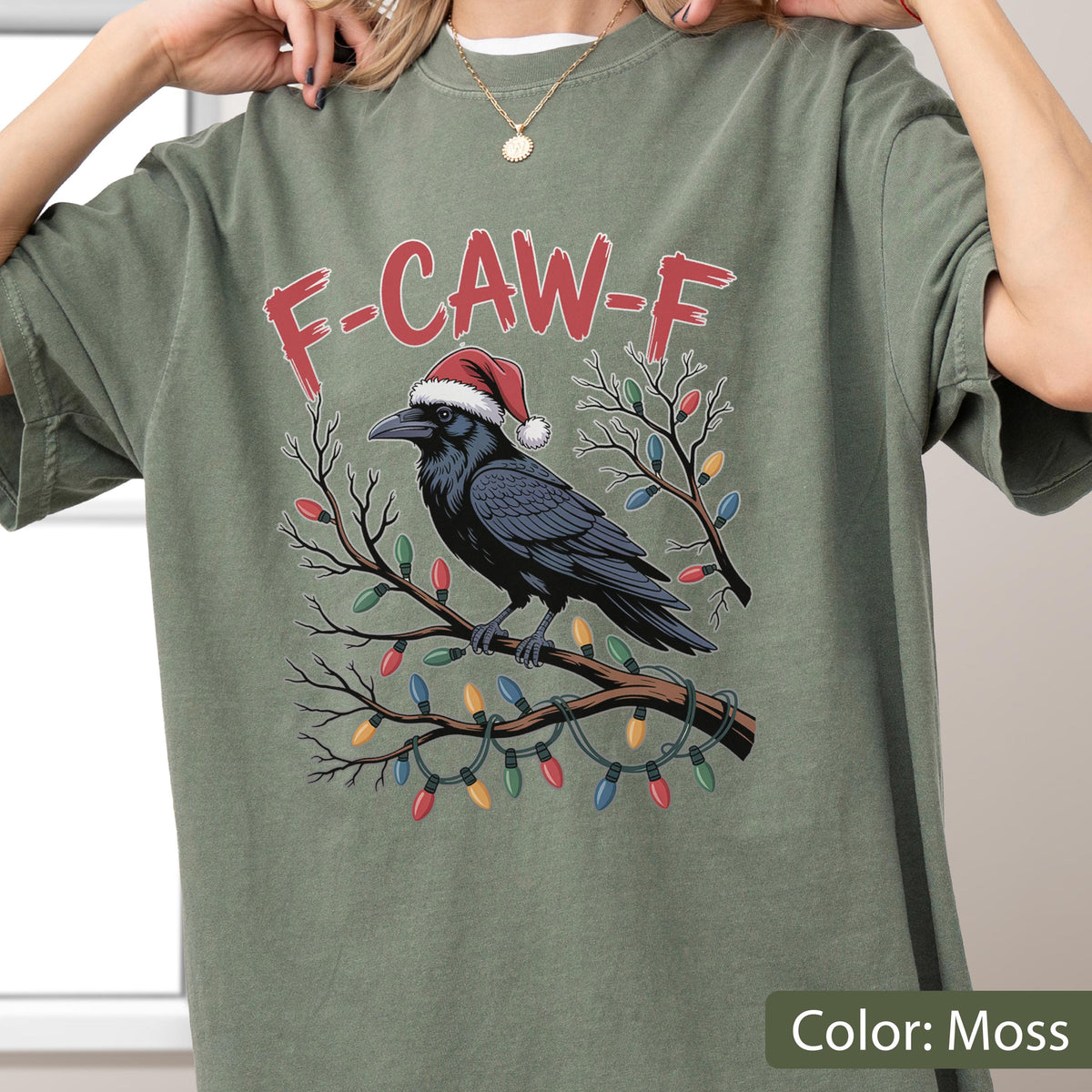 May include: A moss green t-shirt featuring a graphic of a black crow wearing a red and white Santa hat, perched on a bare tree branch wrapped in colorful Christmas lights. The text F-CAW-F is printed in red above the crow in a distressed font.