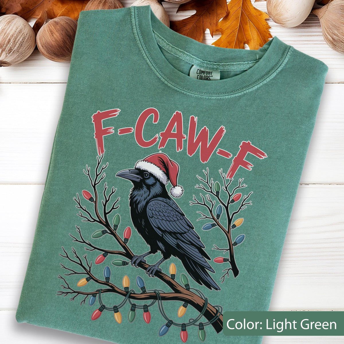 May include: A light green crewneck t-shirt featuring a graphic of a black raven wearing a red and white Santa hat, perched on a brown branch wrapped in colorful Christmas lights. The text F-CAW-F is printed in red above the raven illustration.