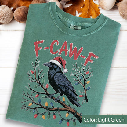 May include: A light green crewneck t-shirt featuring a graphic of a black raven wearing a red and white Santa hat, perched on a brown branch wrapped in colorful Christmas lights. The text F-CAW-F is printed in red above the raven illustration.
