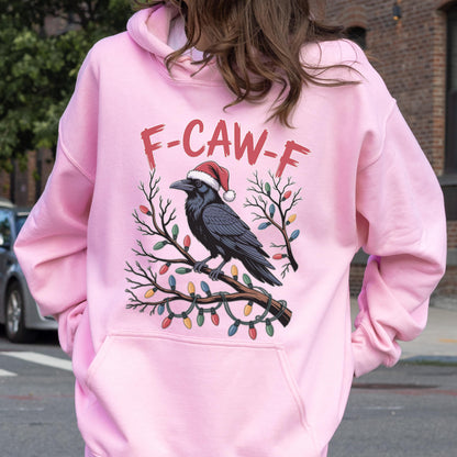 May include: A light pink hooded sweatshirt featuring a graphic of a black crow wearing a red Santa hat, perched on a branch wrapped in colorful Christmas lights. The text F-CAW-F is printed in red above the crow. The hoodie has long sleeves.