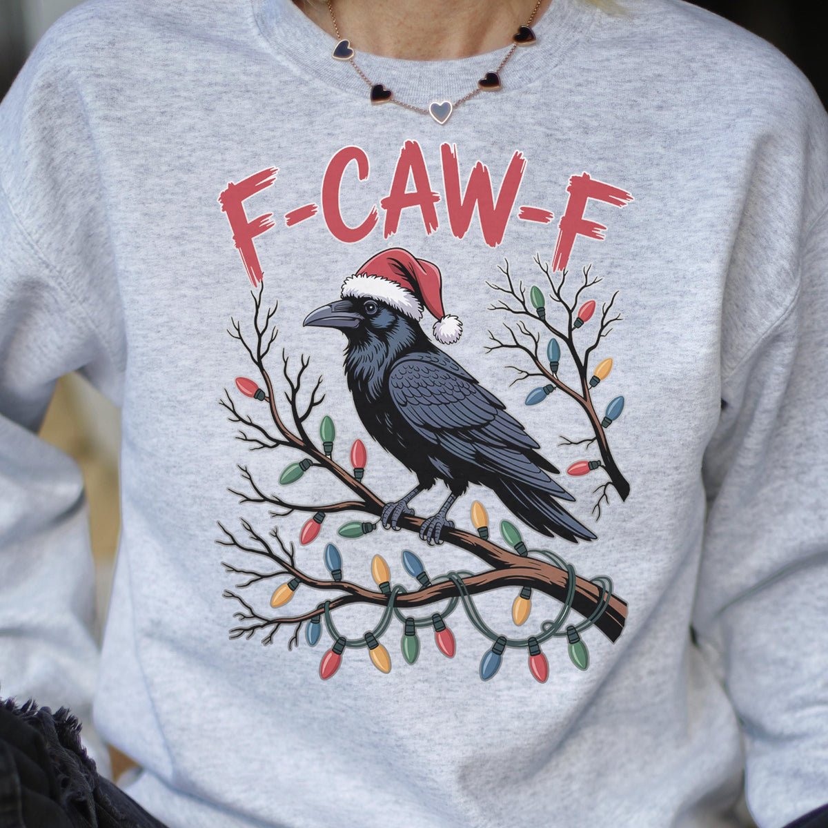 May include: A light gray crewneck sweatshirt featuring a graphic of a black crow wearing a red Santa hat, perched on a bare tree branch wrapped in colorful Christmas lights. The text F-CAW-F is printed in red above the crow illustration.