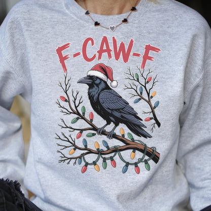 May include: A light gray crewneck sweatshirt featuring a graphic of a black crow wearing a red Santa hat, perched on a bare tree branch wrapped in colorful Christmas lights. The text F-CAW-F is printed in red above the crow illustration.