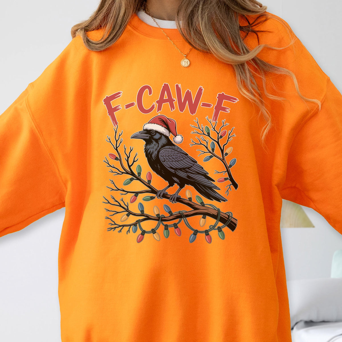 May include: A bright orange crewneck sweatshirt featuring a graphic of a black raven wearing a red and white Santa hat, perched on a branch wrapped in colorful Christmas lights, with the red text F-CAW-F printed in an arched design above.