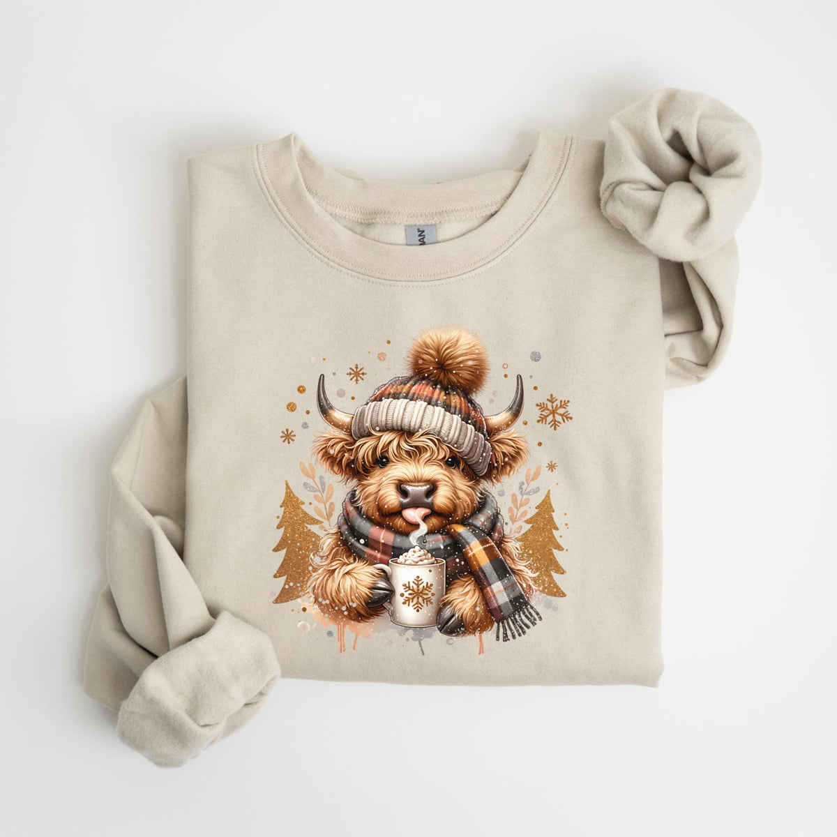 May include: Beige sweatshirt featuring a watercolor illustration of a Highland cow wearing a plaid hat and scarf, holding a mug with a snowflake design. The cow has its tongue out. The design includes snowflakes and gold trees.