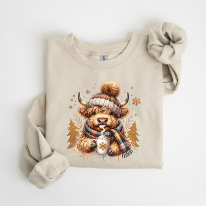 May include: Beige sweatshirt featuring a watercolor illustration of a Highland cow wearing a plaid hat and scarf, holding a mug with a snowflake design. The cow has its tongue out. The design includes snowflakes and gold trees.