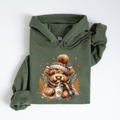 May include: An olive green hoodie featuring a graphic of a Highland cow wearing a plaid hat and scarf, holding a mug of hot cocoa. The cow has a fluffy brown coat and is sticking out its tongue. Gold and white accents complete the winter theme.