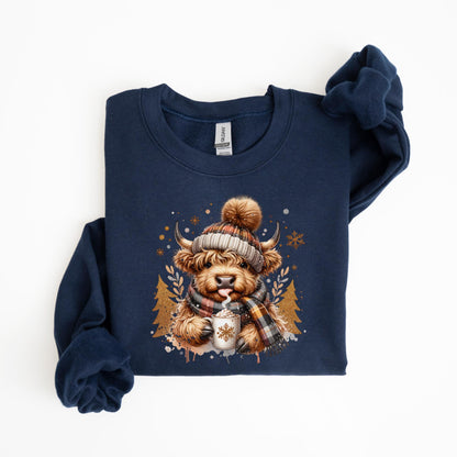 May include: Navy blue sweatshirt featuring a watercolor illustration of a Highland cow wearing a knit hat, scarf, and holding a mug of hot cocoa. The design includes snowflakes and gold-colored trees. The sweatshirt is folded.